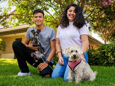 Two people with two small dogs outside in a yard surrounded by a flowering tree