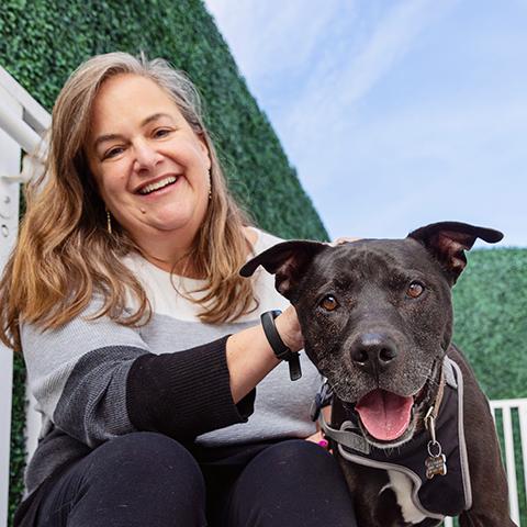 Smiling person next to a smiling black dog with bushes behind them