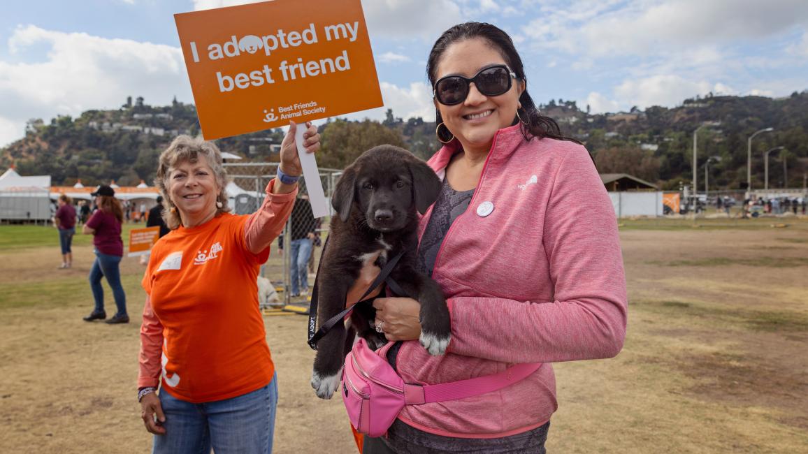 Person holding a puppy at the Los Angeles Super Adoption while another person behind them holds a sign that says I adopted my best friend