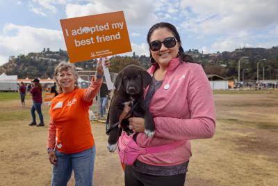 Smiling person holding a puppy with a person behind her holding a sign that says, I adopted my best friend, while at the Los Angeles Super Adoption