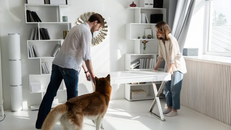 A couple adjusting a desk in a bright, modern living room with a dog observing.