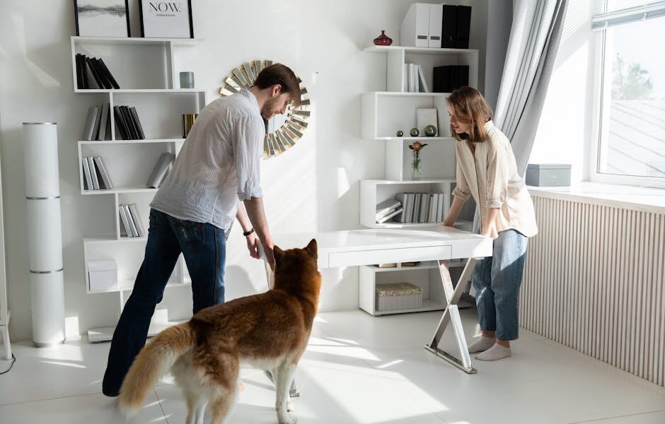 A couple adjusting a desk in a bright, modern living room with a dog observing.