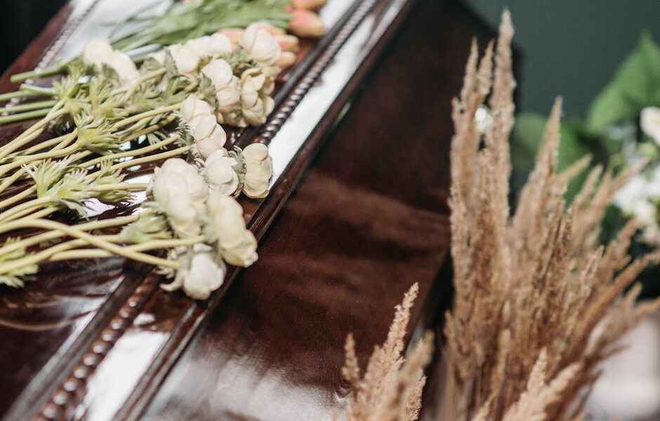 A solemn funeral arrangement featuring a wooden casket adorned with white ranunculus and dried grasses.