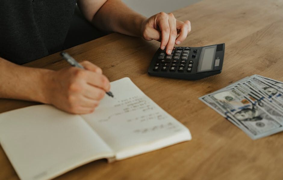 Close-up of hands using a calculator with notebook and cash on a wooden table.