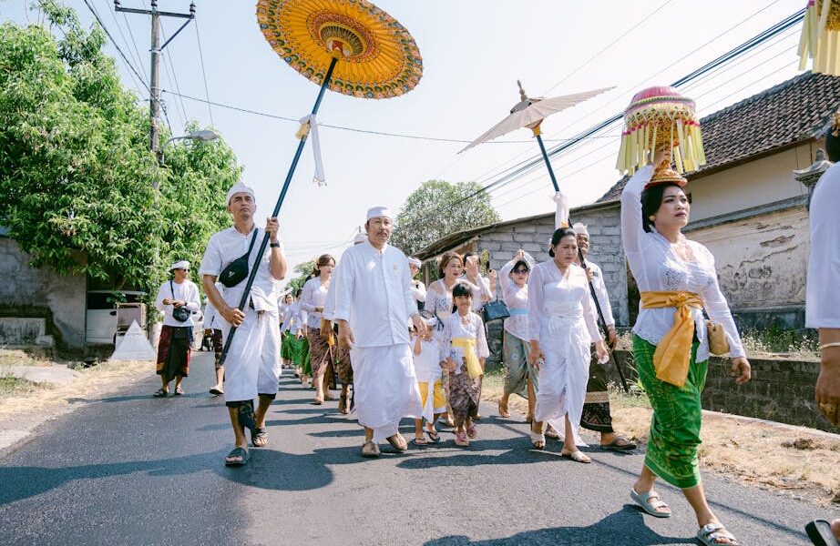 A Balinese procession with traditional attire and vibrant umbrellas on a sunny day.