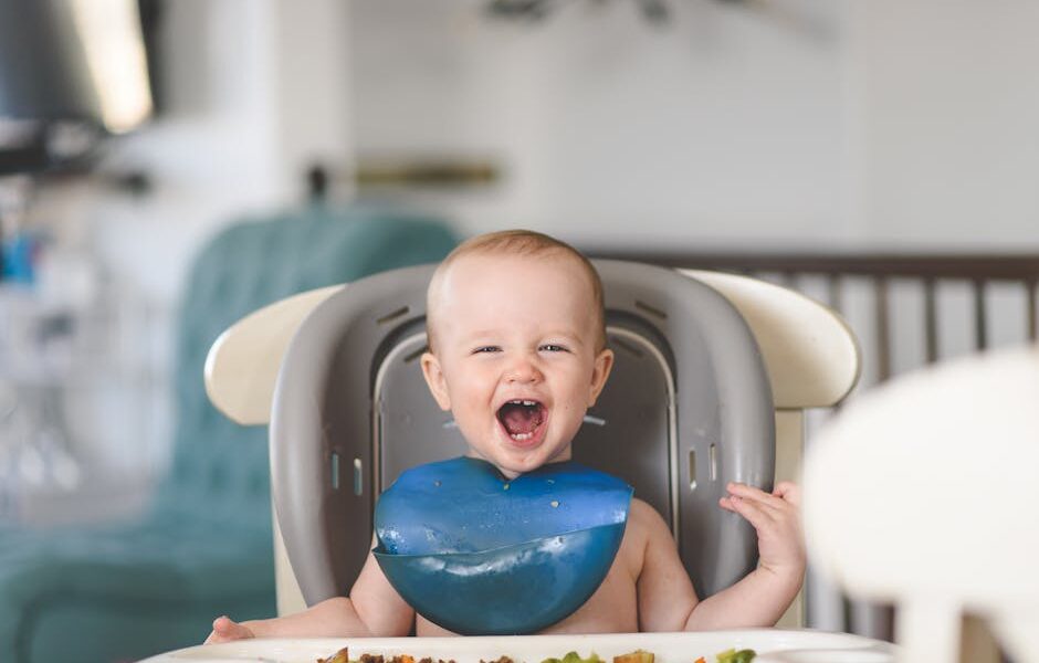 Happy baby laughing in a high chair during mealtime, wearing a blue bib.